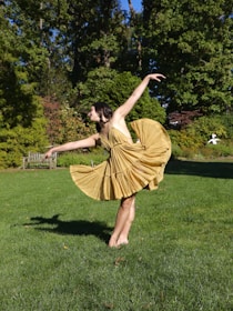 A person is gracefully dancing outdoors on a lush green lawn. The dancer is wearing a flowing yellow dress and has one leg extended while the other is grounded, arms elegantly poised. Surrounding the scene are vibrant green trees and a wooden bench in the background.