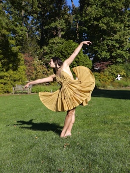 A person is gracefully dancing outdoors on a lush green lawn. The dancer is wearing a flowing yellow dress and has one leg extended while the other is grounded, arms elegantly poised. Surrounding the scene are vibrant green trees and a wooden bench in the background.