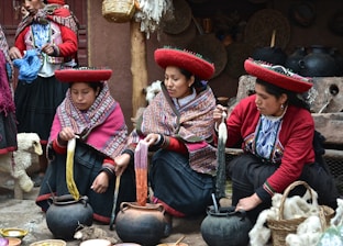 Three women in traditional attire engage in dyeing yarn. They are seated around pots with colorful yarns and appear focused on their work. Their clothing features intricate patterns, and they wear distinctive red hats. Various tools and baskets with yarns surround them, suggesting a weaving or textile-making process.