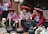 Three women in traditional attire engage in dyeing yarn. They are seated around pots with colorful yarns and appear focused on their work. Their clothing features intricate patterns, and they wear distinctive red hats. Various tools and baskets with yarns surround them, suggesting a weaving or textile-making process.