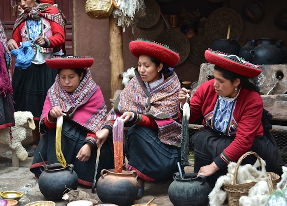 Three women in traditional attire engage in dyeing yarn. They are seated around pots with colorful yarns and appear focused on their work. Their clothing features intricate patterns, and they wear distinctive red hats. Various tools and baskets with yarns surround them, suggesting a weaving or textile-making process.