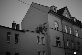 A monochrome photograph of a multi-story residential building with a visible graffiti tag on the side wall. The architecture is typical of urban housing, featuring multiple windows, some with curtains or closed shades. A streetlight is partially captured in the foreground.
