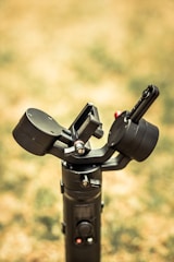 A close-up view of a black camera gimbal set against a blurred outdoor background. The gimbal has a handle with control buttons and a rotating mechanism designed to stabilize cameras.