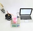 A laboratory setup with a black cylindrical apparatus attached to tubes on the left, a white digital scale displaying '293.2 g' in the center with a transparent beaker containing pink liquid on top of it, a digital thermometer, and a silver-colored laptop with an open screen on the right.