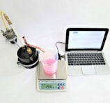 A laboratory setup with a black cylindrical apparatus attached to tubes on the left, a white digital scale displaying '293.2 g' in the center with a transparent beaker containing pink liquid on top of it, a digital thermometer, and a silver-colored laptop with an open screen on the right.