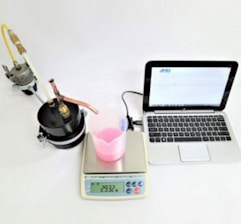 A laboratory setup with a black cylindrical apparatus attached to tubes on the left, a white digital scale displaying '293.2 g' in the center with a transparent beaker containing pink liquid on top of it, a digital thermometer, and a silver-colored laptop with an open screen on the right.