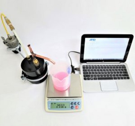 A laboratory setup with a black cylindrical apparatus attached to tubes on the left, a white digital scale displaying '293.2 g' in the center with a transparent beaker containing pink liquid on top of it, a digital thermometer, and a silver-colored laptop with an open screen on the right.