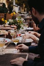 A group of people seated at a dining table outdoors, enjoying a meal together. The table is adorned with a vibrant floral centerpiece featuring yellow and white flowers. Several glasses filled with wine are placed on the table alongside plates of food and appetizers. The background shows blurred greenery and wooden elements, creating a warm and inviting atmosphere.