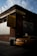 A yellow taxi is parked next to a brick building, partially illuminated by sunlight. There is a sign on the building advertising medallion leases with a $1000 deposit for a brand new car, encouraging people to start today. The scene has a slightly moody atmosphere due to the play of light and shadow, with a partly cloudy sky visible above.