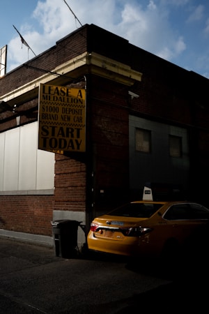 A yellow taxi is parked next to a brick building, partially illuminated by sunlight. There is a sign on the building advertising medallion leases with a $1000 deposit for a brand new car, encouraging people to start today. The scene has a slightly moody atmosphere due to the play of light and shadow, with a partly cloudy sky visible above.