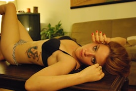 A woman with red hair and a tattoo on her side is lying on a wooden table. She is wearing a black bra and patterned underwear, and has red nail polish. The room has a cozy, dim ambiance with a couch and potted plant in the background.