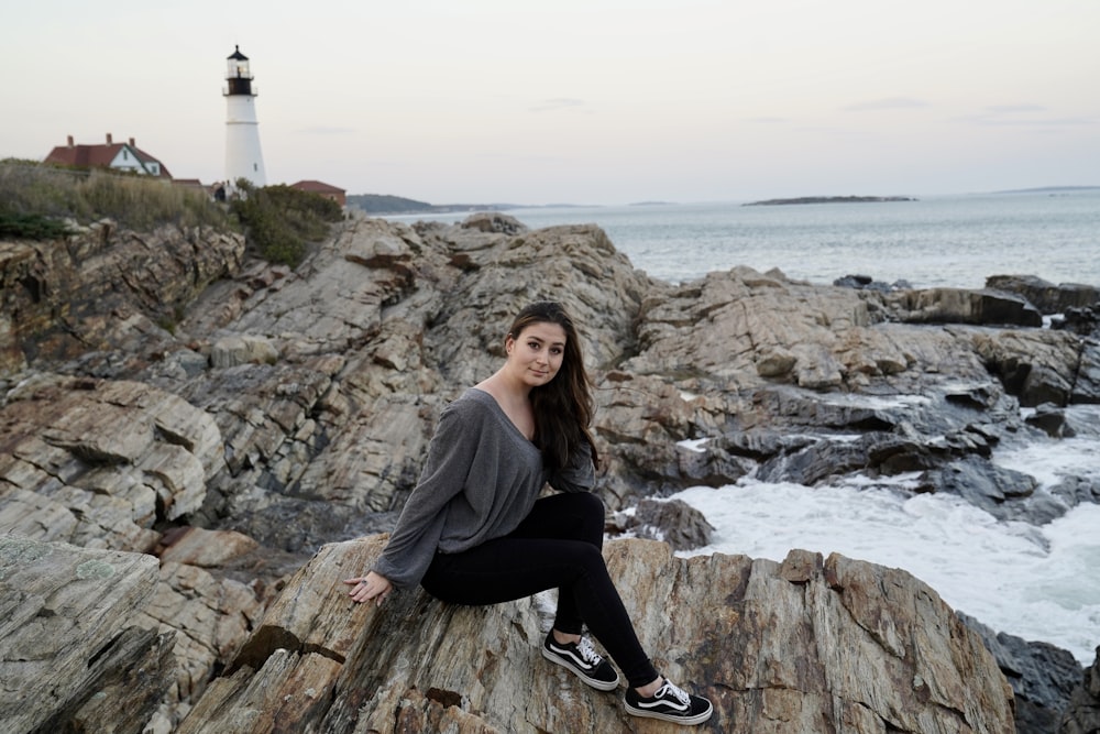 woman in gray sweater and black pants sitting on rock near body of water during daytime