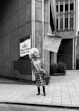 A person is holding a protest sign on a city sidewalk. The sign reads: 'Expose Government KKK Complicity. Legal Help for Protesters' with a phone number. The individual is wearing a patterned dress and a mask, standing in front of a building with signage for a family allergy and asthma center.
