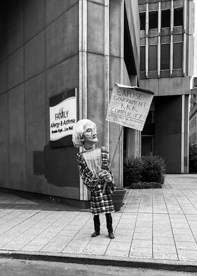A person is holding a protest sign on a city sidewalk. The sign reads: 'Expose Government KKK Complicity. Legal Help for Protesters' with a phone number. The individual is wearing a patterned dress and a mask, standing in front of a building with signage for a family allergy and asthma center.