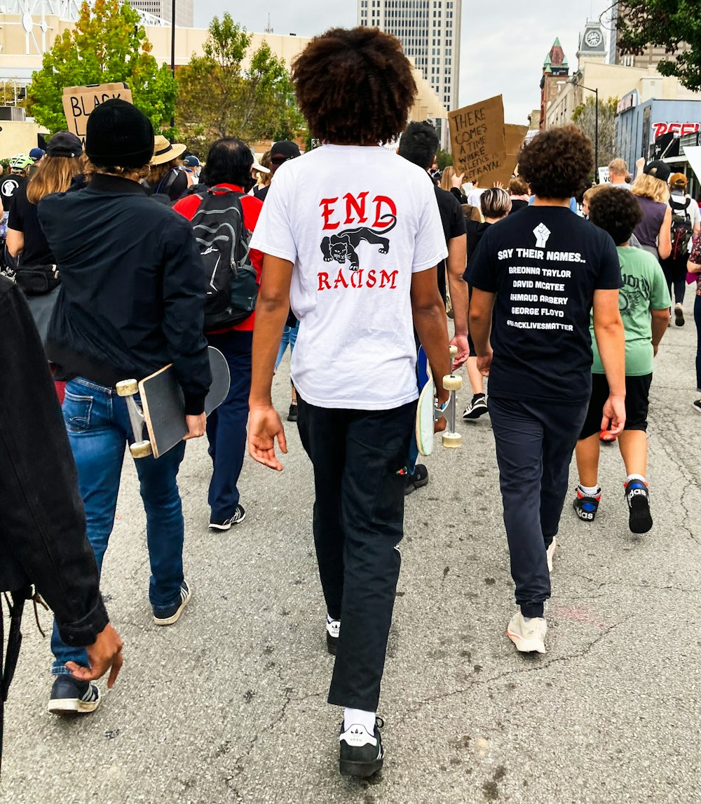 A group of people facing away from the camera, walking in a Black Lives Matter protest in downtown Louisville. Centered is a young Black man with long dark brown curls, wearing a white t-shirt with a Black Panther symbol and red text that reads "End Racism." Another young person to his right wears a black t-shirt with a raised fist and white text that reads "Say Their Names: Breonna Taylor. David McAtee. Ahmaud Arbery. George Floyd. #BlackLivesMatter."