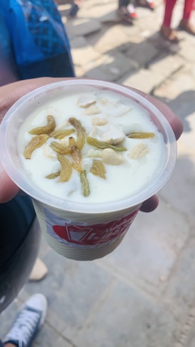 A hand holds a plastic cup filled with a creamy white dessert, garnished with dried fruits and nuts. The background shows a blurred street scene with people walking and the ground is paved with stone tiles.