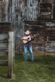 A man wearing a cowboy hat and a plaid shirt is playing an acoustic guitar in front of a rustic wooden barn. The ground is covered with lush green grass, and the barn features weathered wooden planks with a slightly open window.