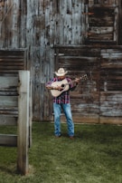 A man wearing a cowboy hat and a plaid shirt is playing an acoustic guitar in front of a rustic wooden barn. The ground is covered with lush green grass, and the barn features weathered wooden planks with a slightly open window.