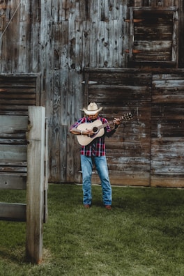 A man wearing a cowboy hat and a plaid shirt is playing an acoustic guitar in front of a rustic wooden barn. The ground is covered with lush green grass, and the barn features weathered wooden planks with a slightly open window.