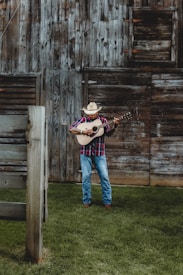 A man wearing a cowboy hat and a plaid shirt is playing an acoustic guitar in front of a rustic wooden barn. The ground is covered with lush green grass, and the barn features weathered wooden planks with a slightly open window.