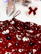 A floral-patterned red fabric is complemented by a delicate necklace. In the background, there are elegant silver high-heeled shoes and a white gift box adorned with a red bow.