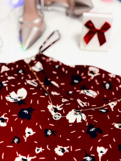 A floral-patterned red fabric is complemented by a delicate necklace. In the background, there are elegant silver high-heeled shoes and a white gift box adorned with a red bow.