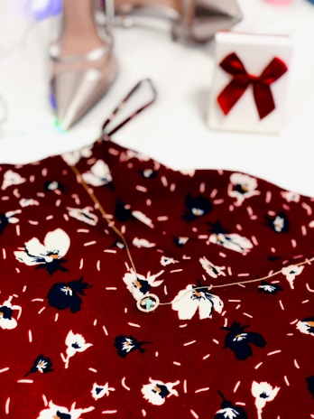 A floral-patterned red fabric is complemented by a delicate necklace. In the background, there are elegant silver high-heeled shoes and a white gift box adorned with a red bow.