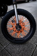A close-up of a motorcycle wheel with striking orange spokes set against a robust black tire. The background consists of paved tiles in a neutral gray, providing a contrasting backdrop to the vibrant wheel components. The metallic suspension fork is prominently visible, shiny and reflective.