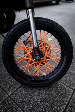 A close-up of a motorcycle wheel with striking orange spokes set against a robust black tire. The background consists of paved tiles in a neutral gray, providing a contrasting backdrop to the vibrant wheel components. The metallic suspension fork is prominently visible, shiny and reflective.