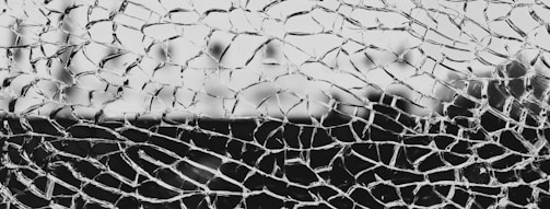 A close-up of shattered glass with jagged, irregular shapes interlocking across the frame. The fragmented glass pieces create a chaotic pattern with a blurred background.