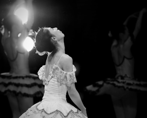 A ballet dancer is captured in a moment of graceful movement, wearing an intricate costume with delicate lace and embellishments. The lighting creates a dramatic contrast, highlighting the dancer's elegant posture and serene expression. In the background, other dancers are partially visible, adding depth to the scene.