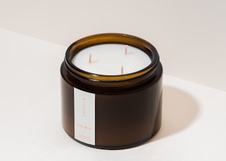A dark amber glass jar containing a white candle with three wicks. The jar features a minimalistic label with text indicating the type of candle.