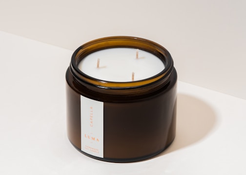 A dark amber glass jar containing a white candle with three wicks. The jar features a minimalistic label with text indicating the type of candle.