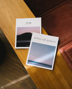 Two booklets titled 'JOB' and 'SONG OF SONGS' are placed on a wooden table. The 'JOB' booklet features a cover image of a silhouette of hills or mountains against a sunset sky. The 'SONG OF SONGS' booklet displays a serene scene of the ocean with a beam of light shining down from the sky. The surface of the table is smooth and has a natural wood texture.