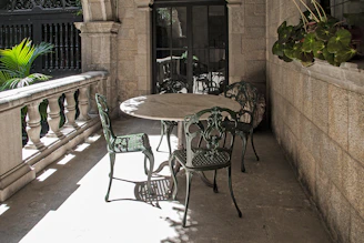An ornate stone balcony features an outdoor setting with a round table and four intricately designed metal chairs. The balcony is framed by a decorative stone railing and lush green plants are visible in the background. There is a touch of sunlight casting shadows on the ground, while potted plants hang on the wall, adding a touch of greenery.