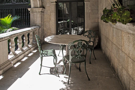An ornate stone balcony features an outdoor setting with a round table and four intricately designed metal chairs. The balcony is framed by a decorative stone railing and lush green plants are visible in the background. There is a touch of sunlight casting shadows on the ground, while potted plants hang on the wall, adding a touch of greenery.