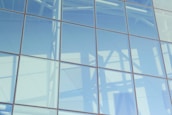 A large glass facade reflecting light and shadows, with a visible grid of metal frames. The reflections include structural elements and soft, diffuse lighting.