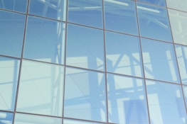 A large glass facade reflecting light and shadows, with a visible grid of metal frames. The reflections include structural elements and soft, diffuse lighting.