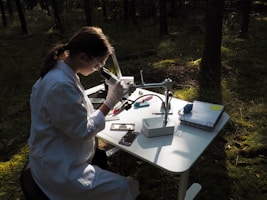 A person wearing a lab coat and protective goggles is sitting at a table in the forest, using a laboratory microscope. Various scientific instruments and a stack of papers are organized on the table, which is illuminated by dappled sunlight filtering through the trees.