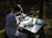 A person wearing a lab coat and protective goggles is sitting at a table in the forest, using a laboratory microscope. Various scientific instruments and a stack of papers are organized on the table, which is illuminated by dappled sunlight filtering through the trees.