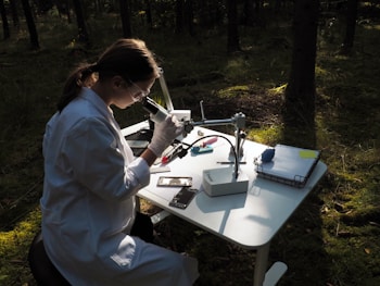 A person wearing a lab coat and protective goggles is sitting at a table in the forest, using a laboratory microscope. Various scientific instruments and a stack of papers are organized on the table, which is illuminated by dappled sunlight filtering through the trees.