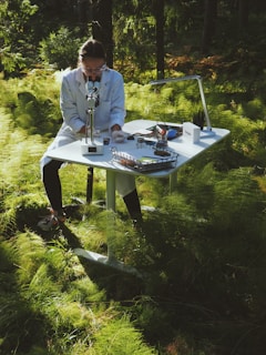 A person wearing a lab coat is sitting at a white desk in a forest, using a microscope. The desk is equipped with various scientific tools and papers. Sunlight filters through the lush green foliage creating a serene and focused atmosphere.