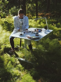 A person wearing a lab coat is sitting at a white desk in a forest, using a microscope. The desk is equipped with various scientific tools and papers. Sunlight filters through the lush green foliage creating a serene and focused atmosphere.