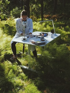 A person wearing a lab coat is sitting at a white desk in a forest, using a microscope. The desk is equipped with various scientific tools and papers. Sunlight filters through the lush green foliage creating a serene and focused atmosphere.
