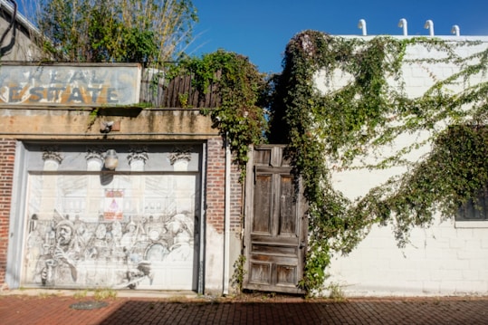 A weathered brick building facade features an artistic mural on its garage door, depicting a busy scene in greyscale. Vines grow over the white wall and the old wooden door in the center, providing a touch of greenery. An old, faded sign above the mural reads 'Real Estate.' The setting is illuminated by sunlight.