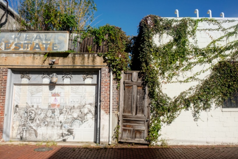 A weathered brick building facade features an artistic mural on its garage door, depicting a busy scene in greyscale. Vines grow over the white wall and the old wooden door in the center, providing a touch of greenery. An old, faded sign above the mural reads 'Real Estate.' The setting is illuminated by sunlight.