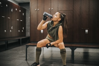 A woman in athletic attire is seated on a bench in a locker room, drinking from a bottle. She wears a sleeveless hoodie, shorts, knee-high socks, and fingerless gloves. The locker room has dark wooden lockers and a tiled floor.