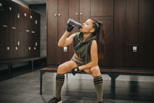 A woman in athletic attire is seated on a bench in a locker room, drinking from a bottle. She wears a sleeveless hoodie, shorts, knee-high socks, and fingerless gloves. The locker room has dark wooden lockers and a tiled floor.