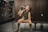 A woman in athletic attire is seated on a bench in a locker room, drinking from a bottle. She wears a sleeveless hoodie, shorts, knee-high socks, and fingerless gloves. The locker room has dark wooden lockers and a tiled floor.