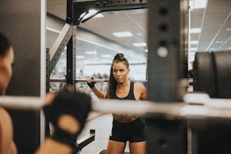 A woman is performing squats in a gym while facing a mirror. She is using a barbell with weights, and appears focused and determined. The gym environment is visible in the background with fitness equipment and fluorescent lighting.